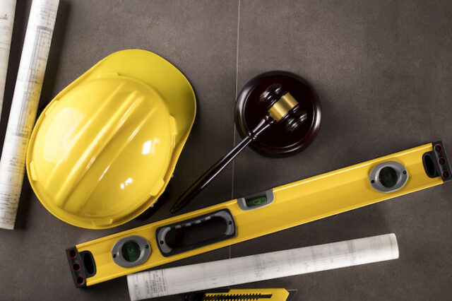 En gul byggehjelm, et gult vatterpas, tegninger og en hammer som man bruger ved brud på byggeregler for tilbygninger ligger på et gråt gulv.