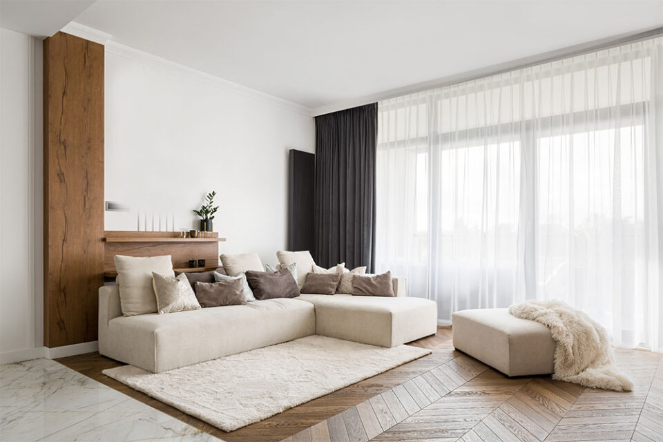 En fin stue med stor sofa og en puf og hvide gardiner som går til gulv